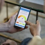 Two people look at a smartphone screen displaying a credit score of 540 out of 715, marked in yellow and orange on a gauge. One person points to the screen. The casual indoor setting hints they might be discussing options like bank accounts for bad credit. | COYYN