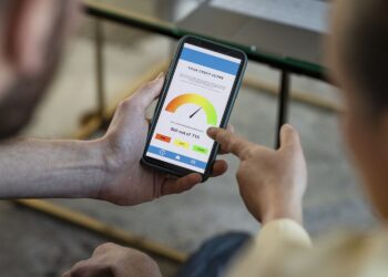Two people look at a smartphone screen displaying a credit score of 540 out of 715, marked in yellow and orange on a gauge. One person points to the screen. The casual indoor setting hints they might be discussing options like bank accounts for bad credit. | COYYN