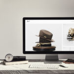 A computer monitor on a desk displays a Squarespace website featuring a beige felt hat with a ribbon and pin. The desk also holds a keyboard, mouse, and notebooks, while the background shows a minimalistic, light-colored wall and a staircase railing. | COYYN