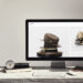 A computer monitor on a desk displays a Squarespace website featuring a beige felt hat with a ribbon and pin. The desk also holds a keyboard, mouse, and notebooks, while the background shows a minimalistic, light-colored wall and a staircase railing. | COYYN