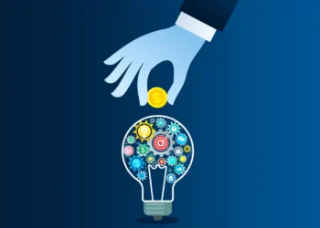 A blue-toned illustration depicts a hand placing a coin into a light bulb filled with gears and mechanical parts. The light bulb symbolizes ideas and innovation, while the coin represents venture capital investment. The background is a gradient of dark blue. | COYYN