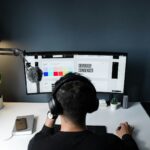 A person wearing headphones sits at a desk, working on a computer with a curved monitor displaying a design program. A microphone is attached to a boom arm, symbolizing their gig economy setup. Nearby are a potted plant, smartphone, and closed laptop. The background is dark gray. | COYYN