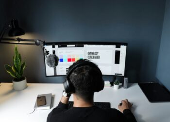 A person wearing headphones sits at a desk, working on a computer with a curved monitor displaying a design program. A microphone is attached to a boom arm, symbolizing their gig economy setup. Nearby are a potted plant, smartphone, and closed laptop. The background is dark gray. | COYYN