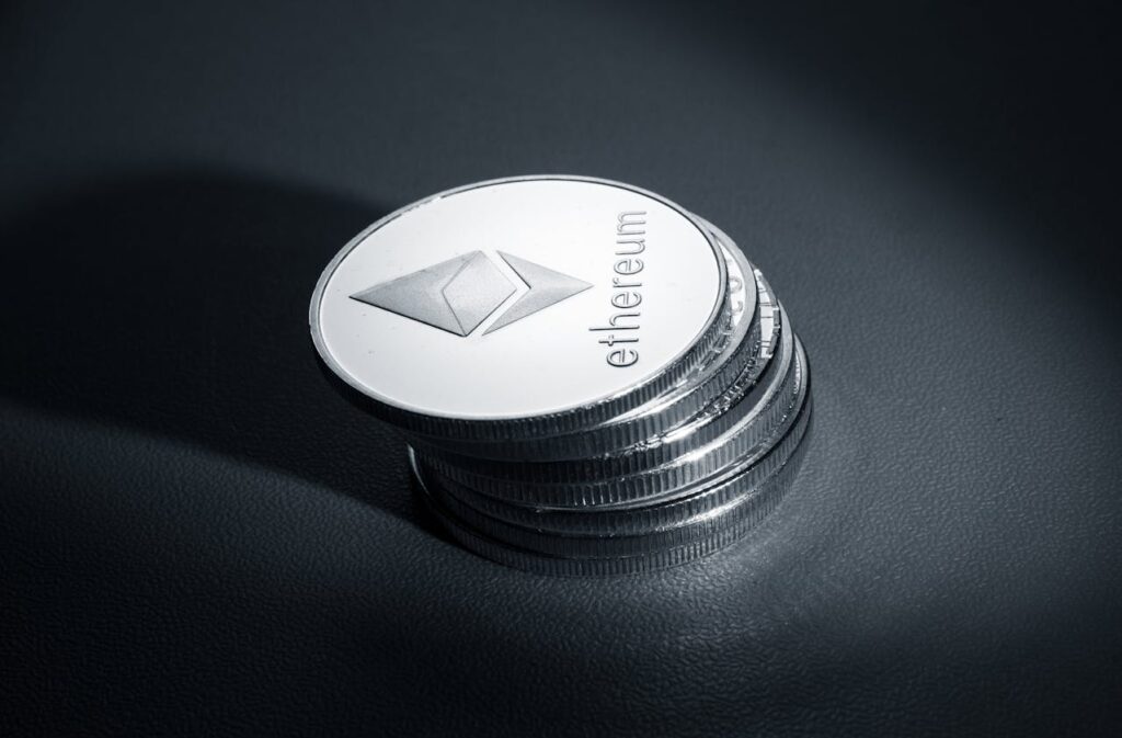 A stack of silver Ethereum Crypto coins, with the top coin displaying the Ethereum logo and the word “ethereum” engraved on it, sits on a dark, smooth surface under a focused light. The polished look captures the essence of crypto sophistication. | COYYN