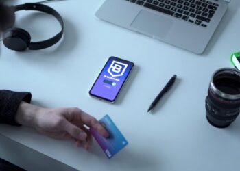 A person holding a blue Visa card sits at a desk with a smartphone displaying a Blockchain app, a laptop, a pair of headphones, a black pen, a book, and a camera lens mug. The desk is white, and the items are neatly arranged. | COYYN