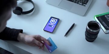 A person holding a blue Visa card sits at a desk with a smartphone displaying a Blockchain app, a laptop, a pair of headphones, a black pen, a book, and a camera lens mug. The desk is white, and the items are neatly arranged. | COYYN