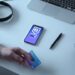 A person holding a blue Visa card sits at a desk with a smartphone displaying a Blockchain app, a laptop, a pair of headphones, a black pen, a book, and a camera lens mug. The desk is white, and the items are neatly arranged. | COYYN