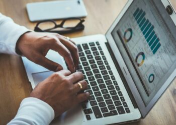 A person is typing on a laptop displaying various data visualizations, including bar charts and pie charts in digital business. The laptop is on a wooden table alongside a smartphone and a pair of glasses. The person is wearing a white long-sleeve shirt and a ring. | COYYN
