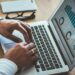 A person is typing on a laptop displaying various data visualizations, including bar charts and pie charts in digital business. The laptop is on a wooden table alongside a smartphone and a pair of glasses. The person is wearing a white long-sleeve shirt and a ring. | COYYN