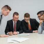 Four men in business attire are gathered around a table looking at documents. Three are dressed in suits and ties, while one wears a shirt and tie without a jacket. They appear to be engaged in a focused discussion about private equity investments or strategies. | COYYN
