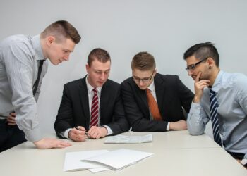 Four men in business attire are gathered around a table looking at documents. Three are dressed in suits and ties, while one wears a shirt and tie without a jacket. They appear to be engaged in a focused discussion about private equity investments or strategies. | COYYN