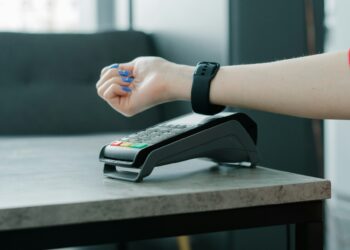 A person's hand, adorned with blue nail polish and a red sleeve, is hovering over a contactless payment terminal on a table, indicating a transaction using digital money. In the background, there's a gray sofa. | COYYN