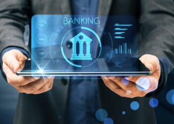 A person in business attire holds a tablet displaying a stylized bank icon and the word "BANKING" in prominent letters. Surrounding the bank icon are various graphical data elements and charts, all glowing with blue accents, symbolizing the future of digital banking. | COYYN
