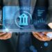 A person in business attire holds a tablet displaying a stylized bank icon and the word "BANKING" in prominent letters. Surrounding the bank icon are various graphical data elements and charts, all glowing with blue accents, symbolizing the future of digital banking. | COYYN