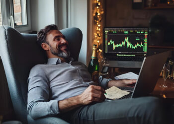 A man sits relaxed in an armchair, smiling at a laptop. Beside him, a computer screen displays stock market charts and the phrase "Best Time to Trade Forex". A champagne bottle and glasses are nearby, indicating celebration or a successful venture. Warm ambient lighting adds to the cozy atmosphere. | COYYN