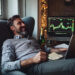 A man sits relaxed in an armchair, smiling at a laptop. Beside him, a computer screen displays stock market charts and the phrase "Best Time to Trade Forex". A champagne bottle and glasses are nearby, indicating celebration or a successful venture. Warm ambient lighting adds to the cozy atmosphere. | COYYN