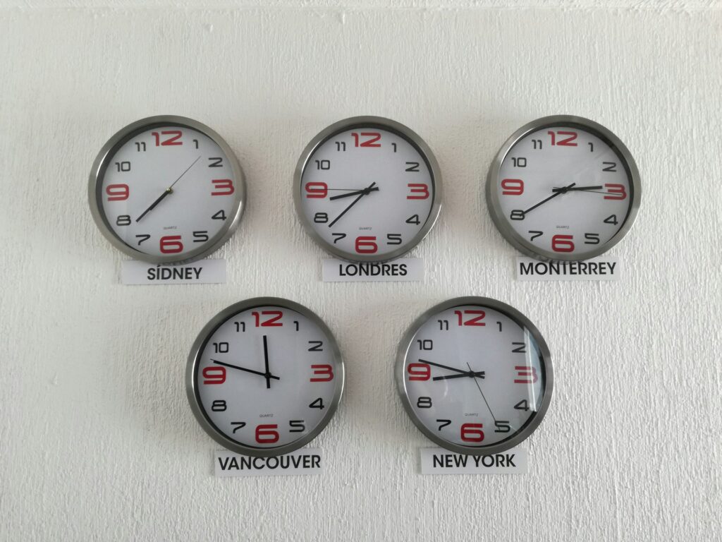 Five wall clocks displaying different times, labeled "SIDNEY," "LONDRES," "MONTERREY," "VANCOUVER," and "NEW YORK," are mounted on a white wall. Each clock shows the current time in its respective city, providing essential insights for anyone involved in forex trading across multiple time zones. | COYYN