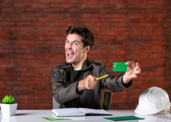 A man cheerfully holds a green debit card, playfully miming cutting it with a pencil. He sits at a desk with an open notebook, a potted plant, and a white hard hat against the red brick wall backdrop—a scene capturing the relief of managing finances for gig worker loans. | COYYN.com