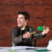 A man cheerfully holds a green debit card, playfully miming cutting it with a pencil. He sits at a desk with an open notebook, a potted plant, and a white hard hat against the red brick wall backdrop—a scene capturing the relief of managing finances for gig worker loans. | COYYN.com