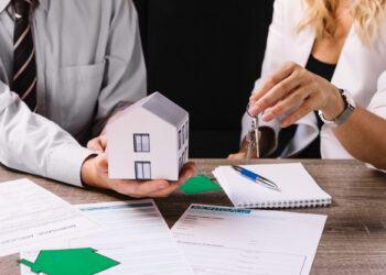 A man holds a model house while a woman hands him a set of keys over a table with mortgage documents, including a crucial mortgage note, contract, and pen. Both are dressed in business attire, indicating a real estate transaction or discussion. | COYYN