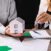 A man holds a model house while a woman hands him a set of keys over a table with mortgage documents, including a crucial mortgage note, contract, and pen. Both are dressed in business attire, indicating a real estate transaction or discussion. | COYYN