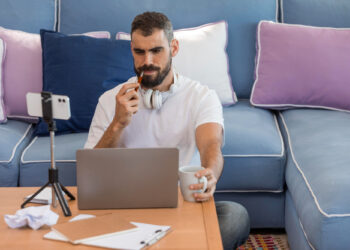 A man with headphones around his neck sits on a sofa with blue and purple cushions, working remotely. With a mug in one hand and a pen in the other, he ponders gig economy jobs. Nearby, a smartphone on a tripod captures it all alongside his notebook and assorted papers. | COYYN.com