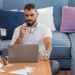 A man with headphones around his neck sits on a sofa with blue and purple cushions, working remotely. With a mug in one hand and a pen in the other, he ponders gig economy jobs. Nearby, a smartphone on a tripod captures it all alongside his notebook and assorted papers. | COYYN.com