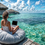 A woman sits on a bean bag on a wooden deck, working on her laptop. Embracing the gig economy lifestyle, she overlooks a crystal-clear turquoise ocean with overwater bungalows visible in the background under a partly cloudy blue sky. | COYYN