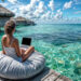 A woman sits on a bean bag on a wooden deck, working on her laptop. Embracing the gig economy lifestyle, she overlooks a crystal-clear turquoise ocean with overwater bungalows visible in the background under a partly cloudy blue sky. | COYYN