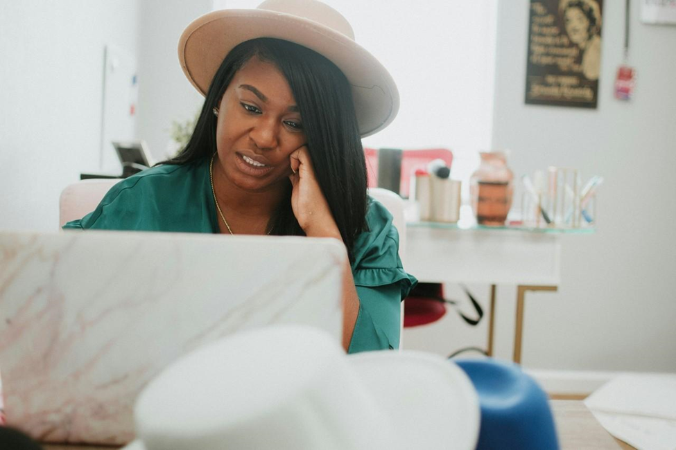 A person in a green top and beige hat is sitting at a desk, looking at a laptop. The room has a light, modern decor with various items and a poster on the wall. | COYYN