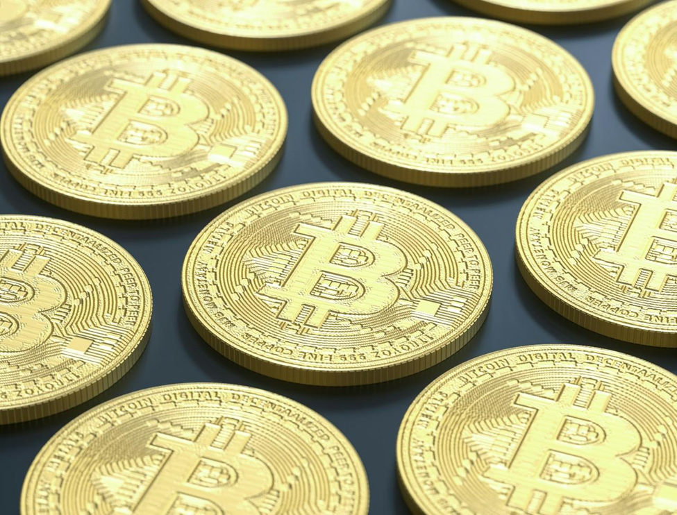 A close-up image of several gold-colored physical Bitcoin coins arranged in rows. Each coin displays the Bitcoin symbol (₿) prominently in the center, with intricate circuit-like patterns around the edges. The background is dark. | COYYN