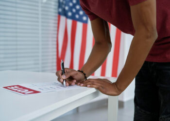 A person in a red shirt leans over a table to fill out a voting form with a pen, ensuring voting security. An American flag is visible in the background, and the word "VOTE" is printed on the paper. | COYYN