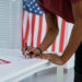 A person in a red shirt leans over a table to fill out a voting form with a pen, ensuring voting security. An American flag is visible in the background, and the word "VOTE" is printed on the paper. | COYYN