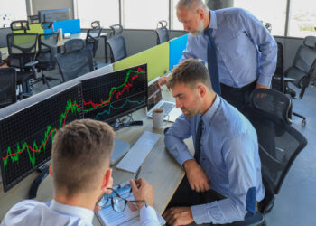 Three men in business attire are intently analyzing stocks and options on financial graphs displayed across multiple computer screens in an office environment with several workstations. One is seated with a focused expression, another points at the screen, and the third observes. | COYYN