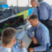 Three men in business attire are intently analyzing stocks and options on financial graphs displayed across multiple computer screens in an office environment with several workstations. One is seated with a focused expression, another points at the screen, and the third observes. | COYYN