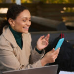 A person, likely engaged in the gig economy, sits outdoors on a bench, wearing a tan coat and green turtleneck, smiling at a smartphone. A laptop is open in front of them amidst bags and a notebook, perfectly capturing the essence of mobile work life. | COYYN