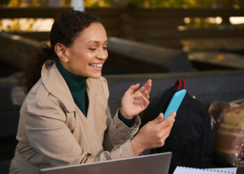 A person, likely engaged in the gig economy, sits outdoors on a bench, wearing a tan coat and green turtleneck, smiling at a smartphone. A laptop is open in front of them amidst bags and a notebook, perfectly capturing the essence of mobile work life. | COYYN