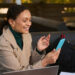 A person, likely engaged in the gig economy, sits outdoors on a bench, wearing a tan coat and green turtleneck, smiling at a smartphone. A laptop is open in front of them amidst bags and a notebook, perfectly capturing the essence of mobile work life. | COYYN