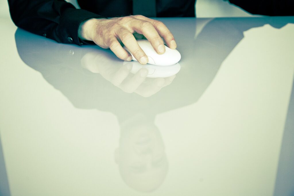 A person wearing a black shirt uses a computer mouse on a highly reflective surface, which captures the reflection of their hand and partially their face. The setting appears minimalistic and modern with a soft, muted color palette. | COYYN