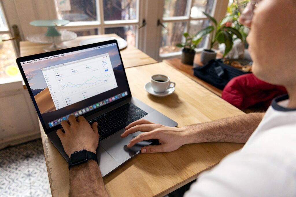 A person is using a laptop at a wooden table, analyzing graphs and data displayed on the screen. A cup of coffee sits nearby. The setting is a bright room with plants by the window. | COYYN