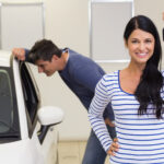 A woman in a striped shirt smiles and holds a car key, having just chosen the right car, in front of a white vehicle. A man in a blue shirt leans down to look inside, both clearly at ease as they explore options at the bustling dealership. | COYYN