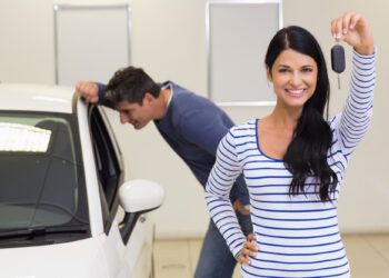 A woman in a striped shirt smiles and holds a car key, having just chosen the right car, in front of a white vehicle. A man in a blue shirt leans down to look inside, both clearly at ease as they explore options at the bustling dealership. | COYYN
