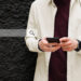 A person wearing a light-colored shirt and a maroon top is holding a smartphone in front of a textured black wall. A translucent search bar graphic with a magnifying glass icon—hinting at the "Find Anyone Online" digital tool for quick searches—overlaps the image on the left. | COYYN