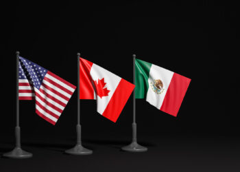 Three small flags on poles against a dark background: from left to right, the flags of the United States, Canada, and Mexico. Each flag is displayed at an angle, standing resilient despite issues like Trump tariffs affecting trade among these neighboring nations. | COYYN