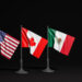 Three small flags on poles against a dark background: from left to right, the flags of the United States, Canada, and Mexico. Each flag is displayed at an angle, standing resilient despite issues like Trump tariffs affecting trade among these neighboring nations. | COYYN