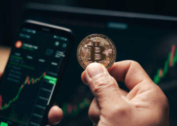 A person holds a physical Bitcoin coin in front of a smartphone displaying a cryptocurrency trading app, highlighting their bitcoin investment. Charts and rates are visible, with a computer screen in the background showing similar data and an encouraging green upward trend. | COYYN