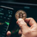 A person holds a physical Bitcoin coin in front of a smartphone displaying a cryptocurrency trading app, highlighting their bitcoin investment. Charts and rates are visible, with a computer screen in the background showing similar data and an encouraging green upward trend. | COYYN