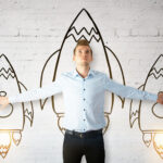 An entrepreneur in a light blue shirt stands with arms outstretched, aligning his hands with two drawn rockets on a white brick wall. A larger central rocket is behind him, appearing to lift off around him. | COYYN