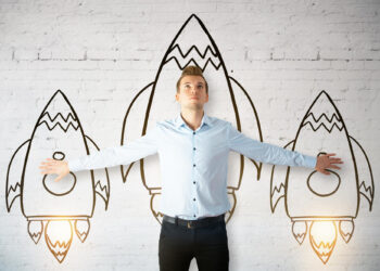 An entrepreneur in a light blue shirt stands with arms outstretched, aligning his hands with two drawn rockets on a white brick wall. A larger central rocket is behind him, appearing to lift off around him. | COYYN