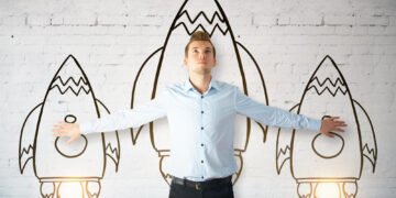 An entrepreneur in a light blue shirt stands with arms outstretched, aligning his hands with two drawn rockets on a white brick wall. A larger central rocket is behind him, appearing to lift off around him. | COYYN